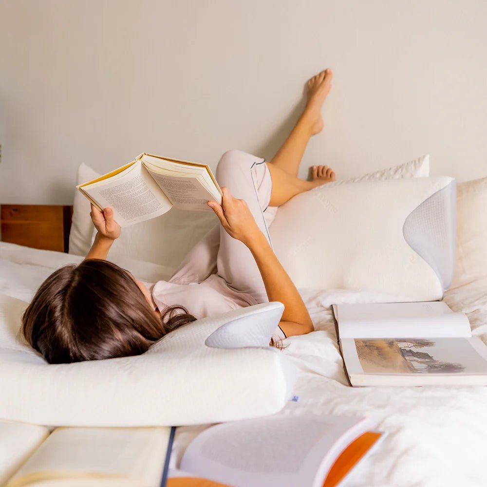 Kobieta leżąca i czytająca na łóżku z biało-szarą poduszką ergonomiczną.